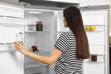 Young woman near empty refrigerator in kitchen, back view