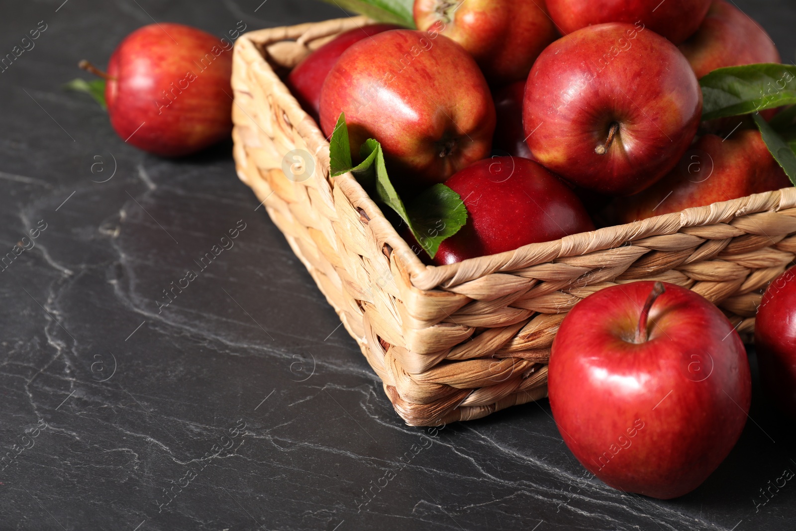 Photo of Fresh red apples and leaves in basket on dark grey table. Space for text