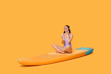 Photo of Happy woman with refreshing drink talking on smartphone on SUP board against orange background