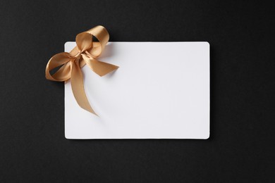 Photo of Blank gift card with golden bow on black background, top view. Space for text