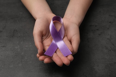 Woman holding purple awareness ribbon on grey background, closeup