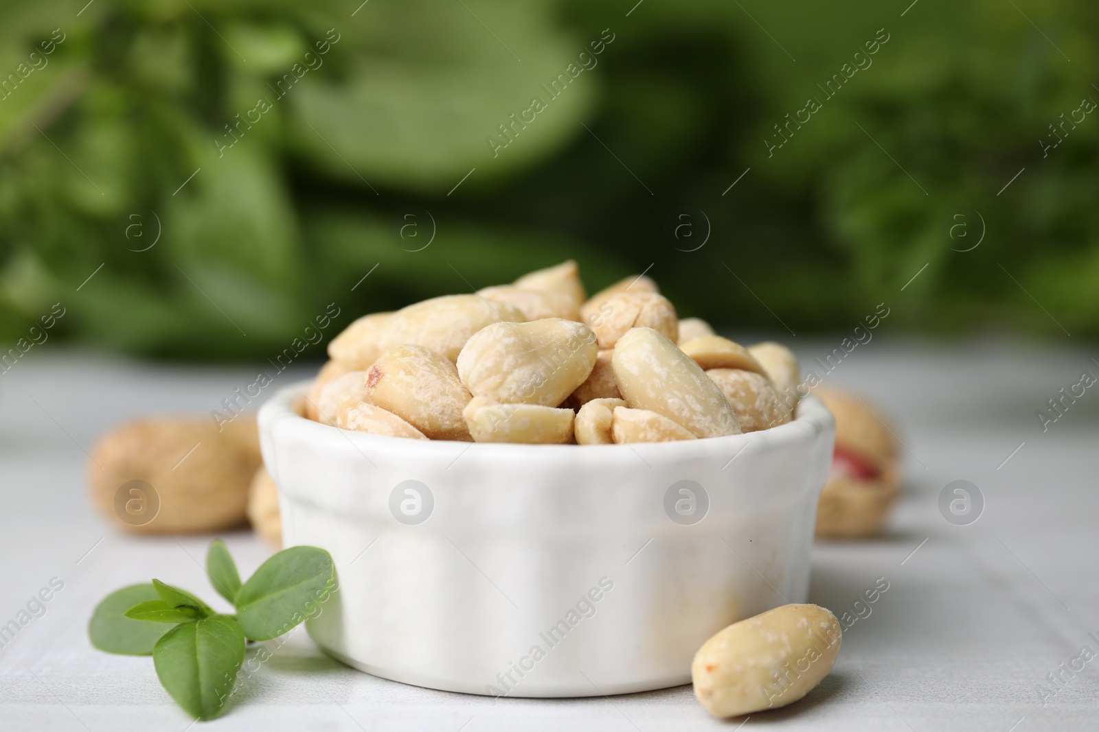 Photo of Fresh peeled peanuts in bowl on white tiled table against blurred green background, closeup