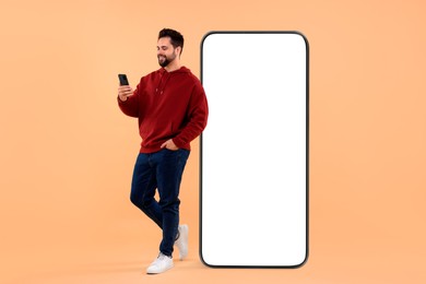 Image of Man with mobile phone standing near huge device with empty screen on dark beige background. Mockup for design