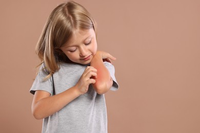 Photo of Suffering from allergy. Little girl scratching her arm on light brown background, space for text