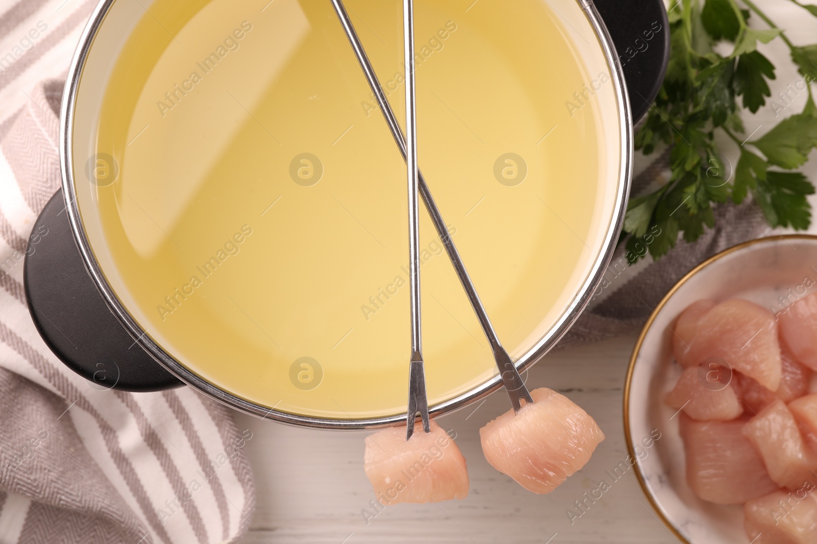 Photo of Fondue pot with oil, forks, raw meat pieces and parsley on white wooden table, flat lay