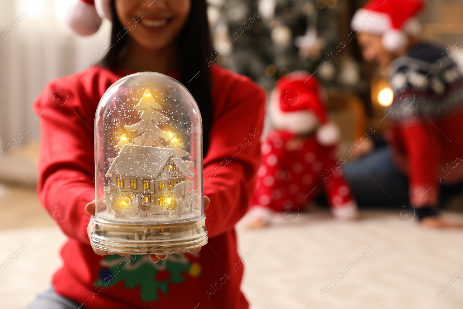 Photo of Woman in red Christmas sweater holding decorative snow globe at home, closeup