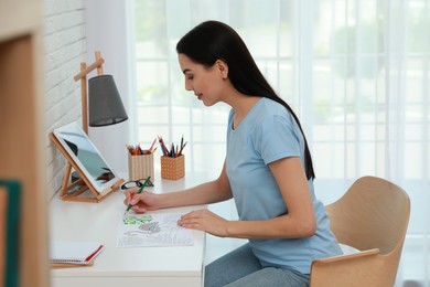 Photo of Young woman coloring antistress page at desk indoors