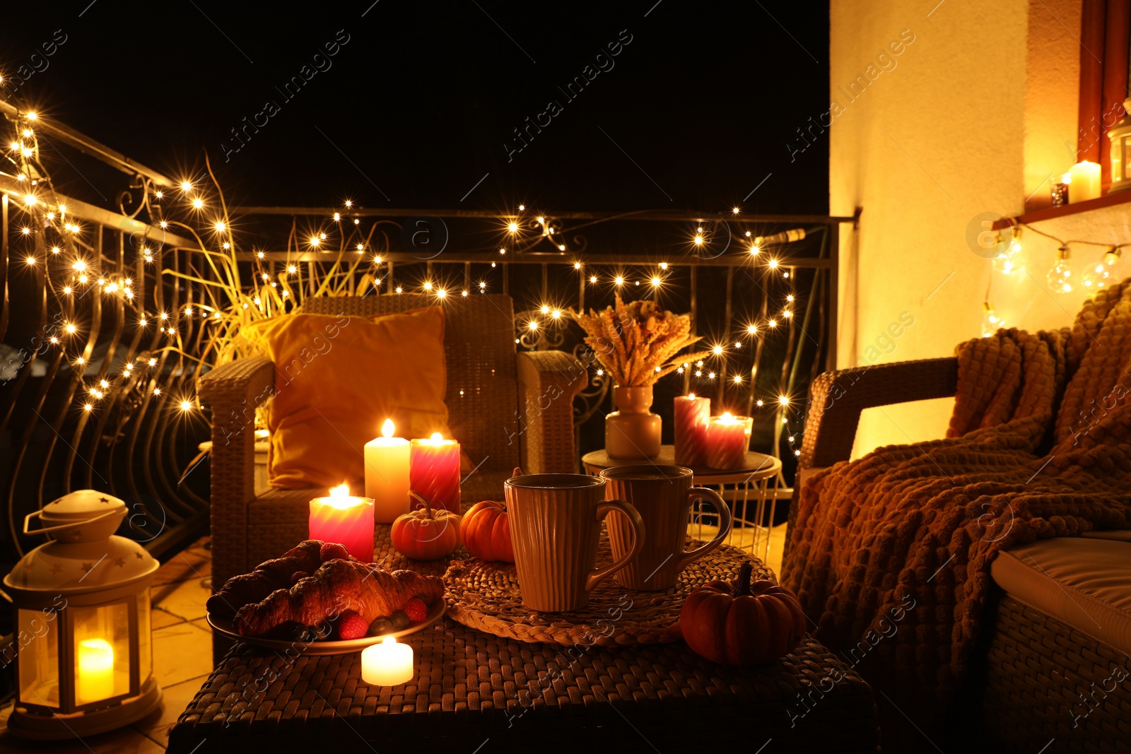 Photo of Rattan furniture, cups, fairy lights, burning candles and other autumn decor on outdoor terrace at night