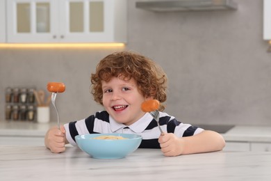 Photo of Cute little boy holding forks with sausages and bowl of pasta at table in kitchen