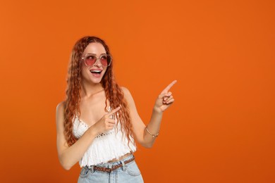 Photo of Stylish young hippie woman in sunglasses pointing at something on orange background, space for text