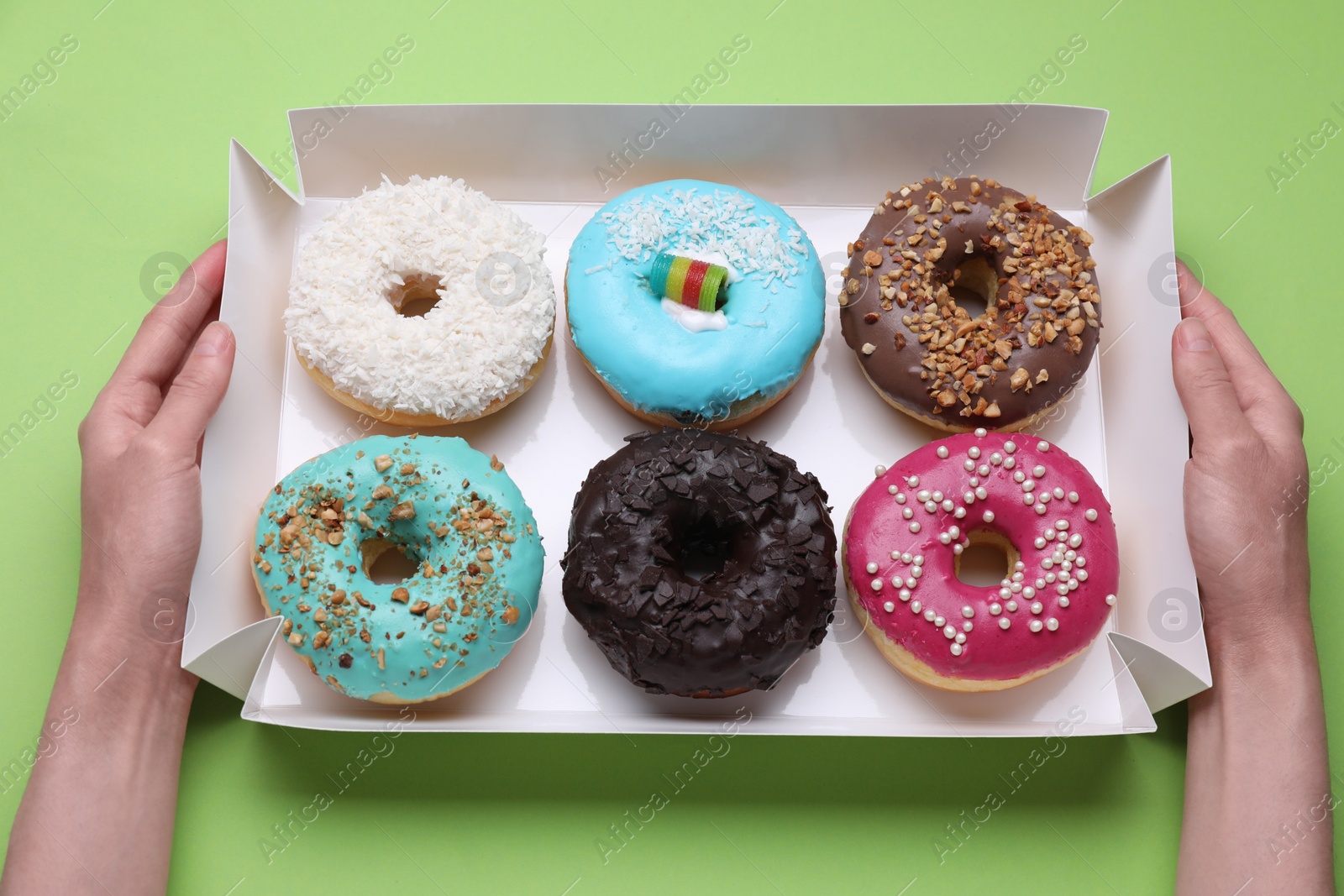 Photo of Woman holding box with tasty glazed donuts on light green background, top view