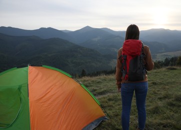 Woman enjoying mountain landscape near camping tent at sunset, back view