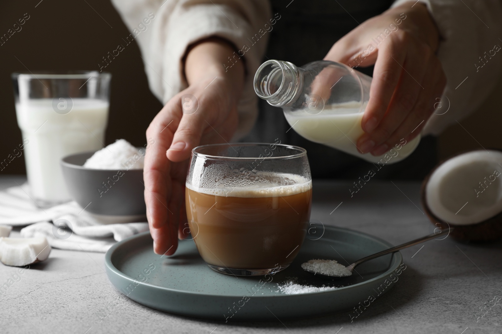 Photo of Woman holding bottle with coconut milk near glass of coffee at light grey table, closeup