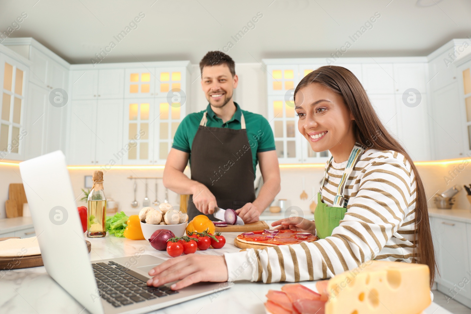 Photo of Happy couple making pizza together while watching online cooking course via laptop in kitchen