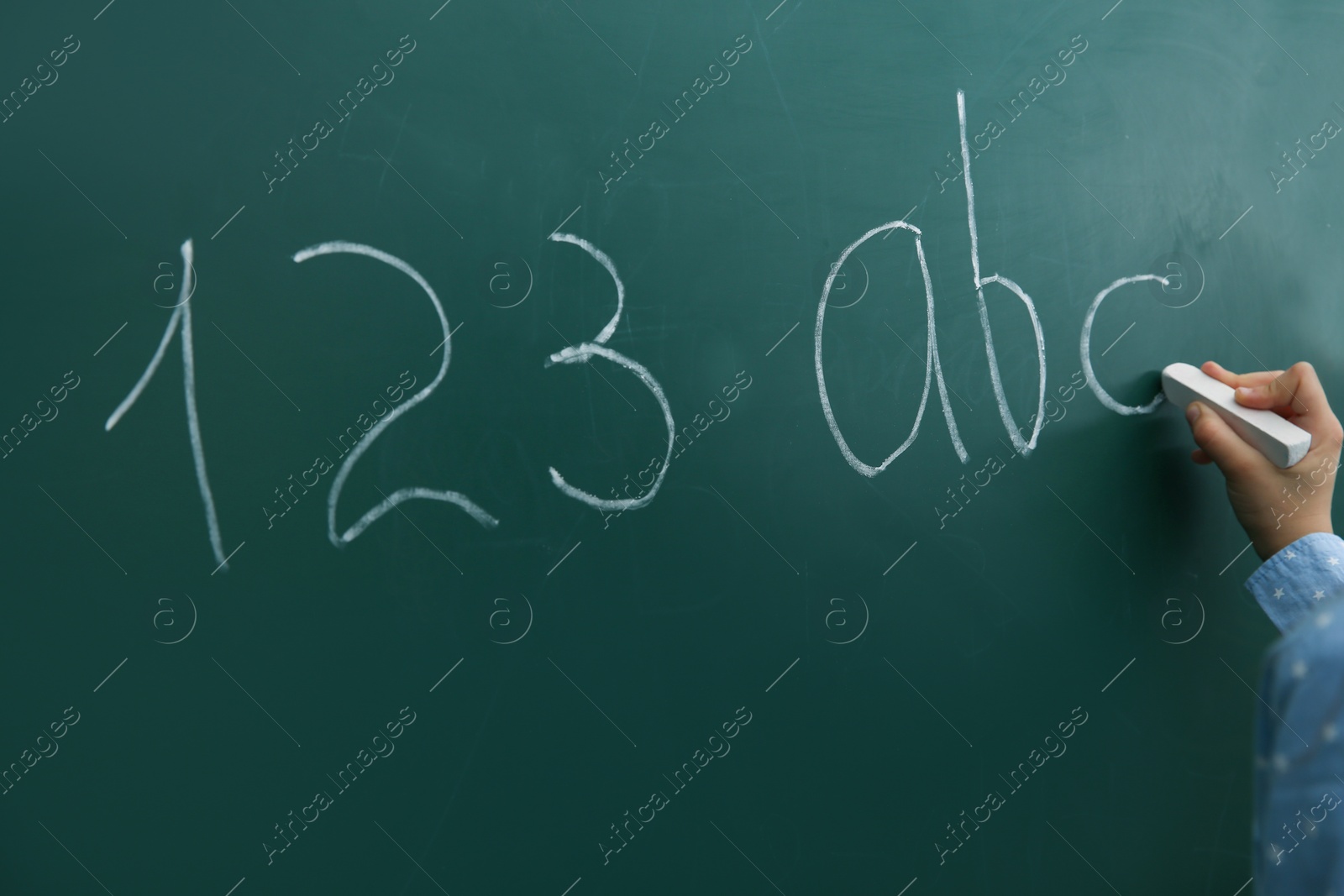 Photo of Little child writing letters and numbers on chalkboard, closeup