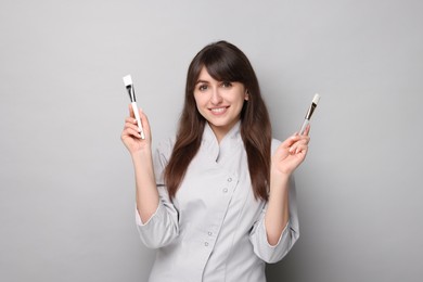 Cosmetologist with cosmetic brushes on grey background