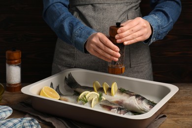Photo of Woman grinding pepper onto raw sea bass fish with lemon and rosemary at wooden table, closeup