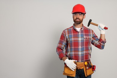 Professional builder in hard hat with hammer and tool belt on light background, space for text