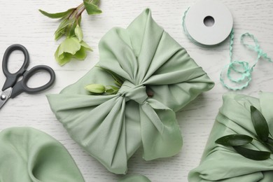 Photo of Furoshiki technique. Gifts packed in green fabrics, hellebore flowers, scissors and decorative ribbon on white wooden table, flat lay