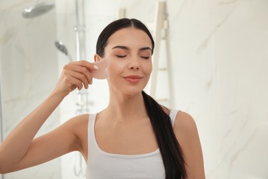 Photo of Beautiful young woman doing facial massage with gua sha tool in bathroom