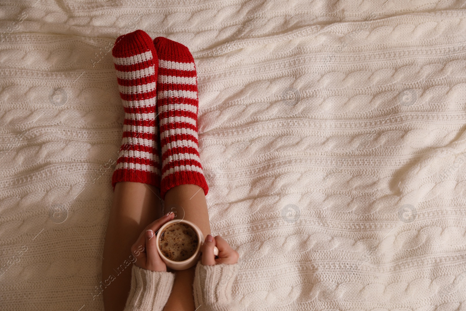 Photo of Woman in warm socks with cup of hot drink relaxing on knitted blanket, top view. Space for text