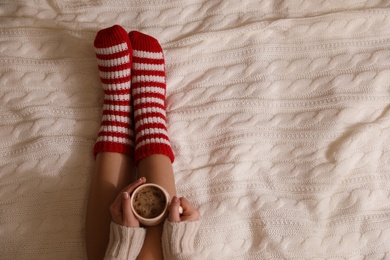 Woman in warm socks with cup of hot drink relaxing on knitted blanket, top view. Space for text