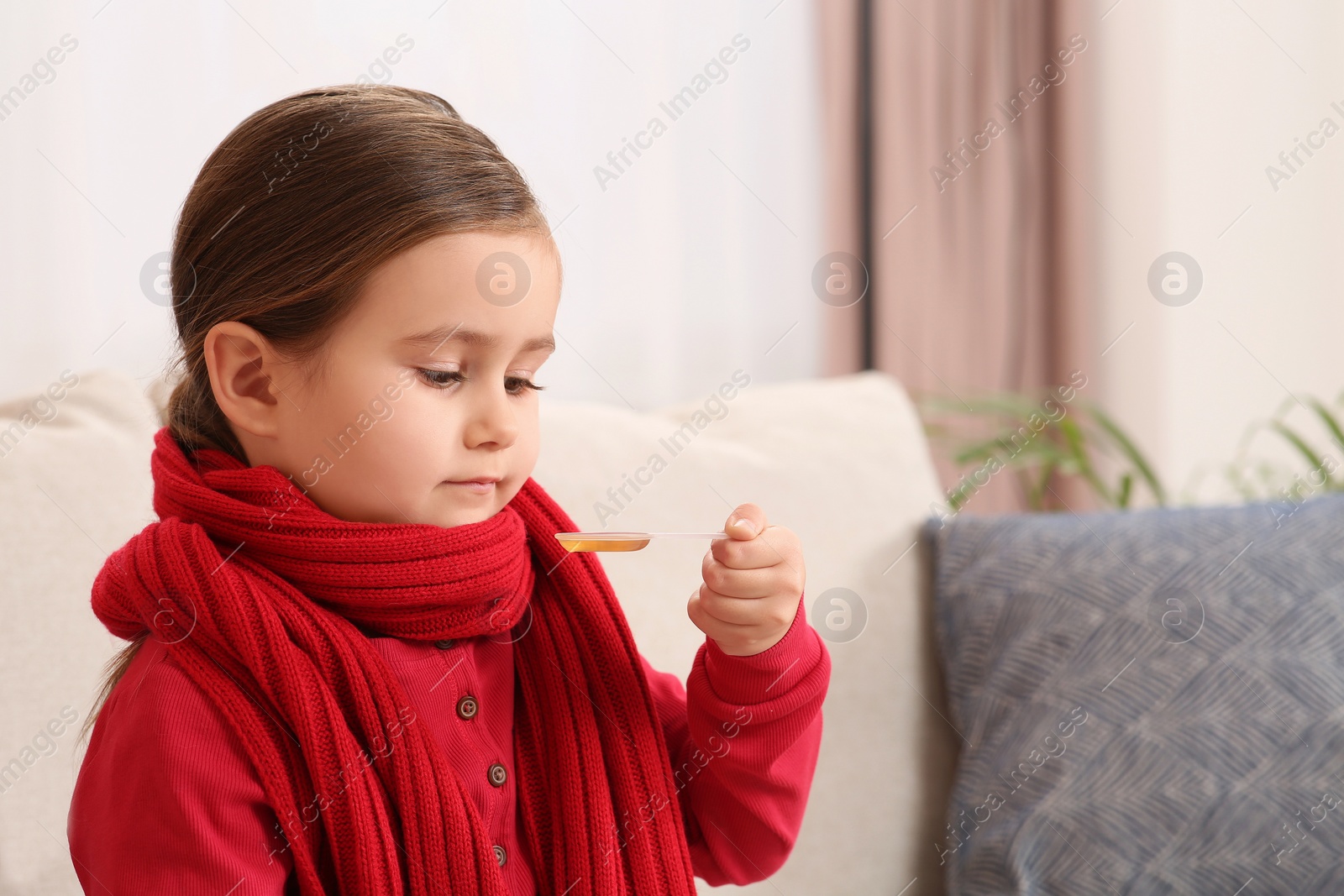 Photo of Cute girl holding measuring spoon with cough syrup on sofa indoors. Effective medicine