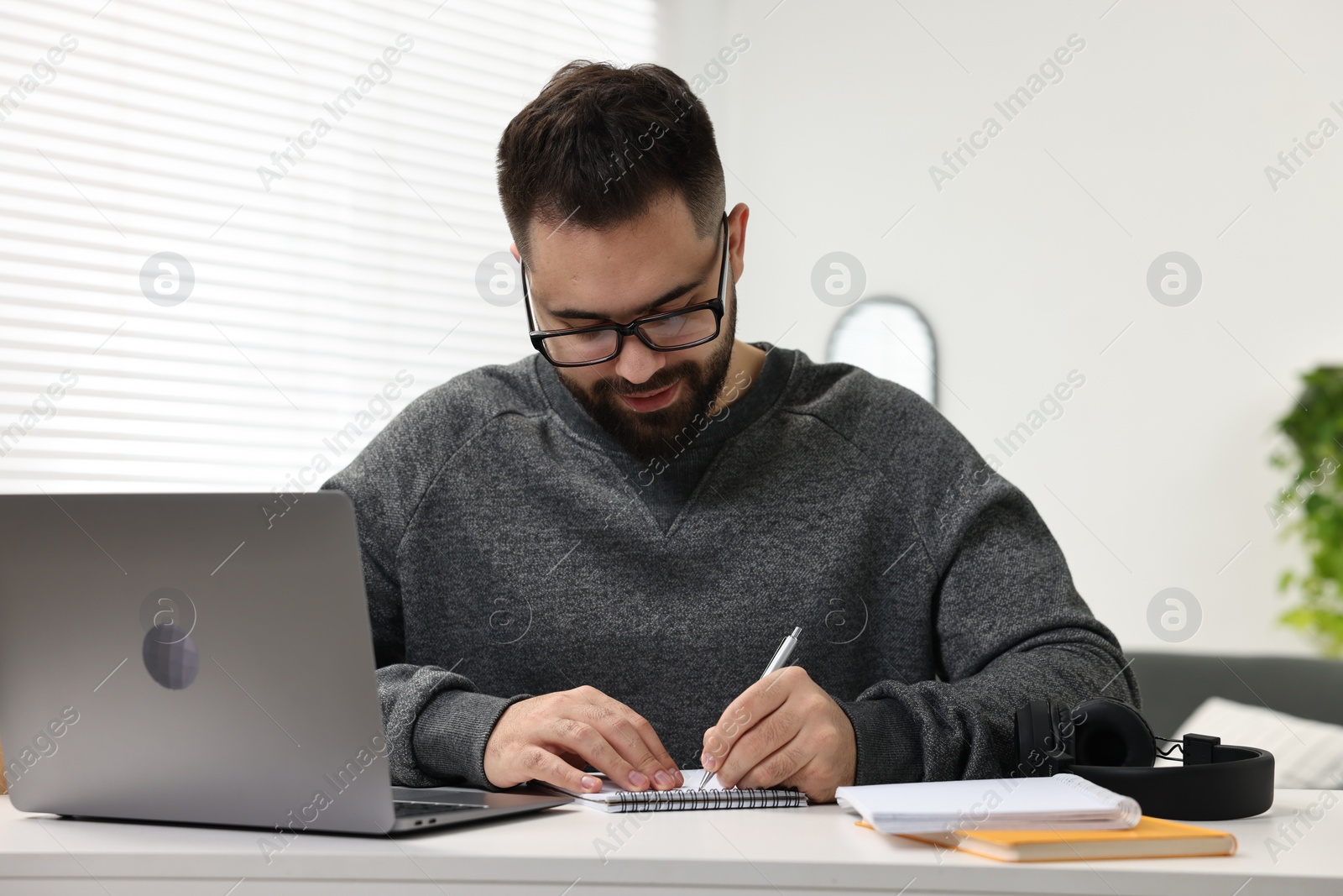 Photo of E-learning. Young man taking notes during online lesson at white table indoors