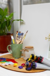 Canvas, wooden artist's palette with colorful paints and brushes on white table near window