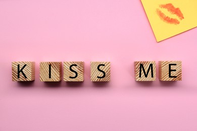 Photo of Wooden cubes with phrase Kiss Me and sticky note with lipstick mark on pink background, flat lay