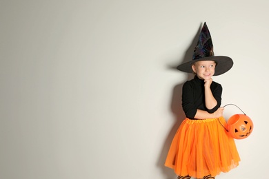 Photo of Cute little girl with pumpkin candy bucket wearing Halloween costume on light background. Space for text