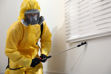 Pest control worker in protective suit spraying insecticide on window sill indoors