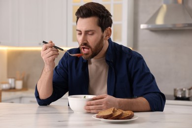 Man eating delicious tomato soup at light marble table in kitchen
