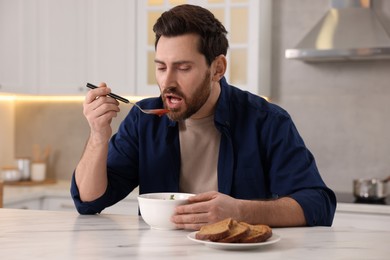 Photo of Man eating delicious tomato soup at light marble table in kitchen