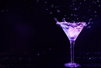 Photo of Glass of martini with ice and splashes in neon lights on dark background. Space for text