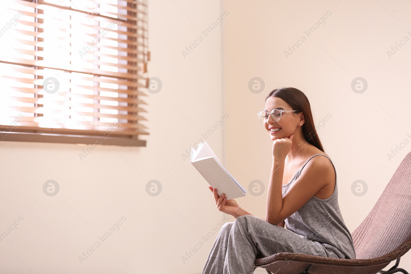 Photo of Young woman reading book near window with blinds at home. Space for text