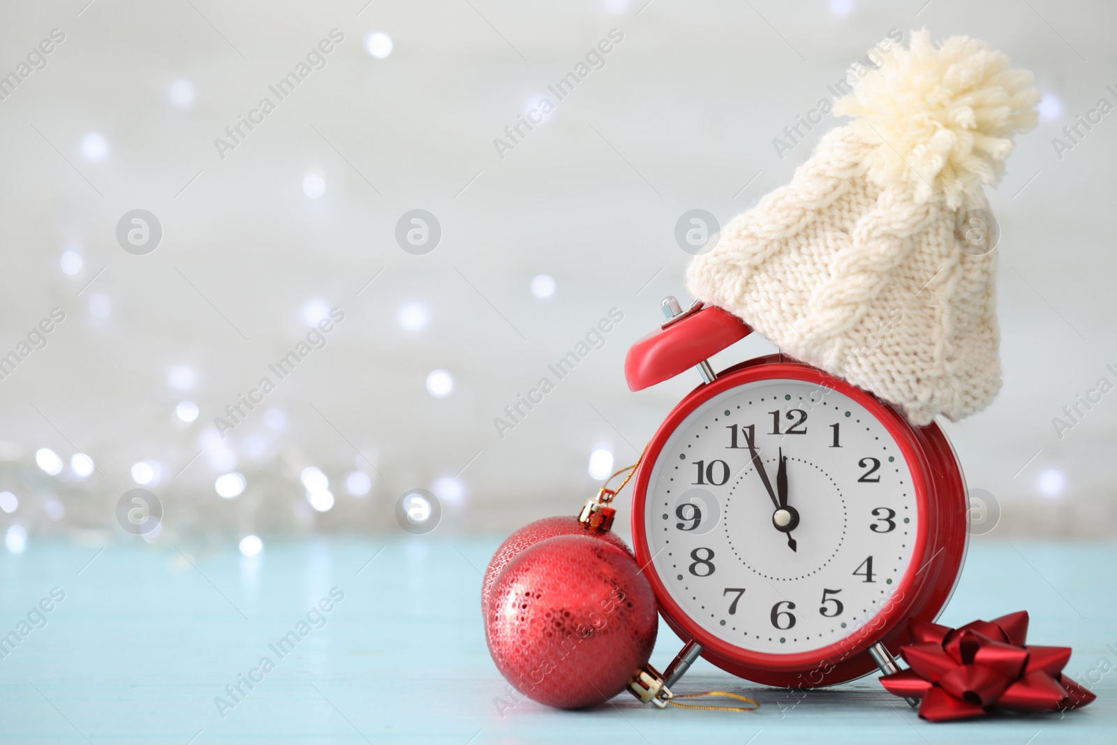 Photo of Alarm clock with decor on light blue table against blurred Christmas lights, space for text. New Year countdown