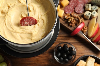 Photo of Fork with piece of sausage, melted cheese in fondue pot and other products on wooden table, flat lay