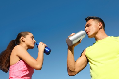 Young sporty couple drinking from water bottles against blue sky on sunny day