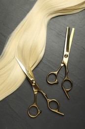 Photo of Professional hairdresser scissors with blonde hair strand on dark grey table, flat lay