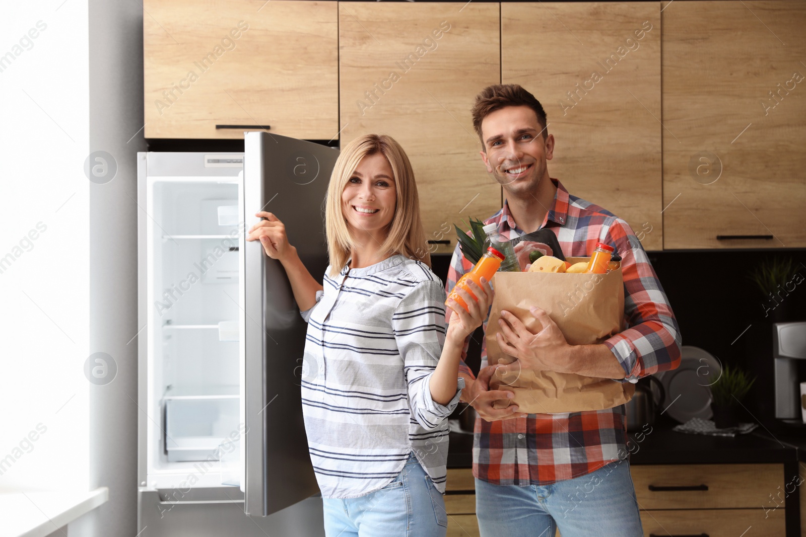 Photo of Happy couple with paper bag full of products near refrigerator in kitchen