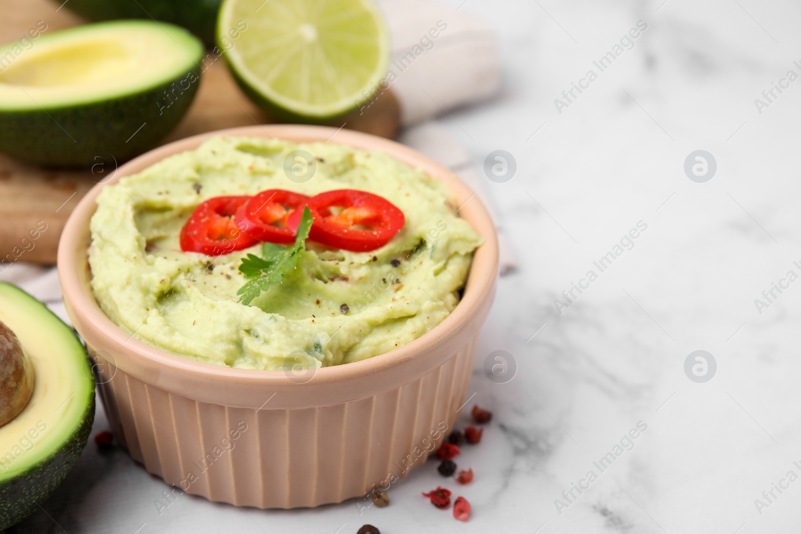 Photo of Bowl of delicious guacamole and ingredients on white marble table, closeup. Space for text