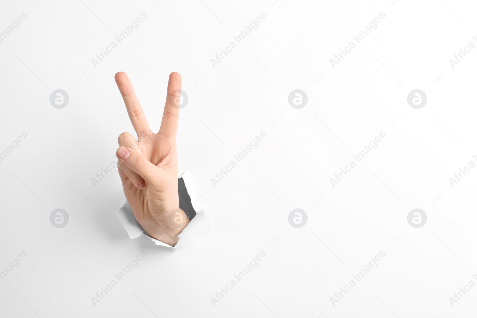 Photo of Woman showing peace sign through hole in torn white paper, space for text. Victory concept