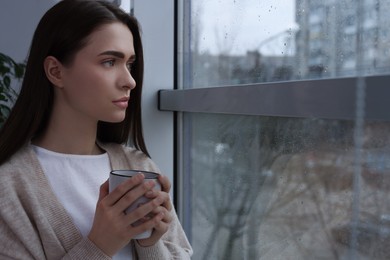 Photo of Melancholic young woman with drink looking out of window on rainy day, space for text. Loneliness concept