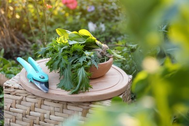 Photo of Wooden board with fresh green herbs and pruner on wicker basket outdoors