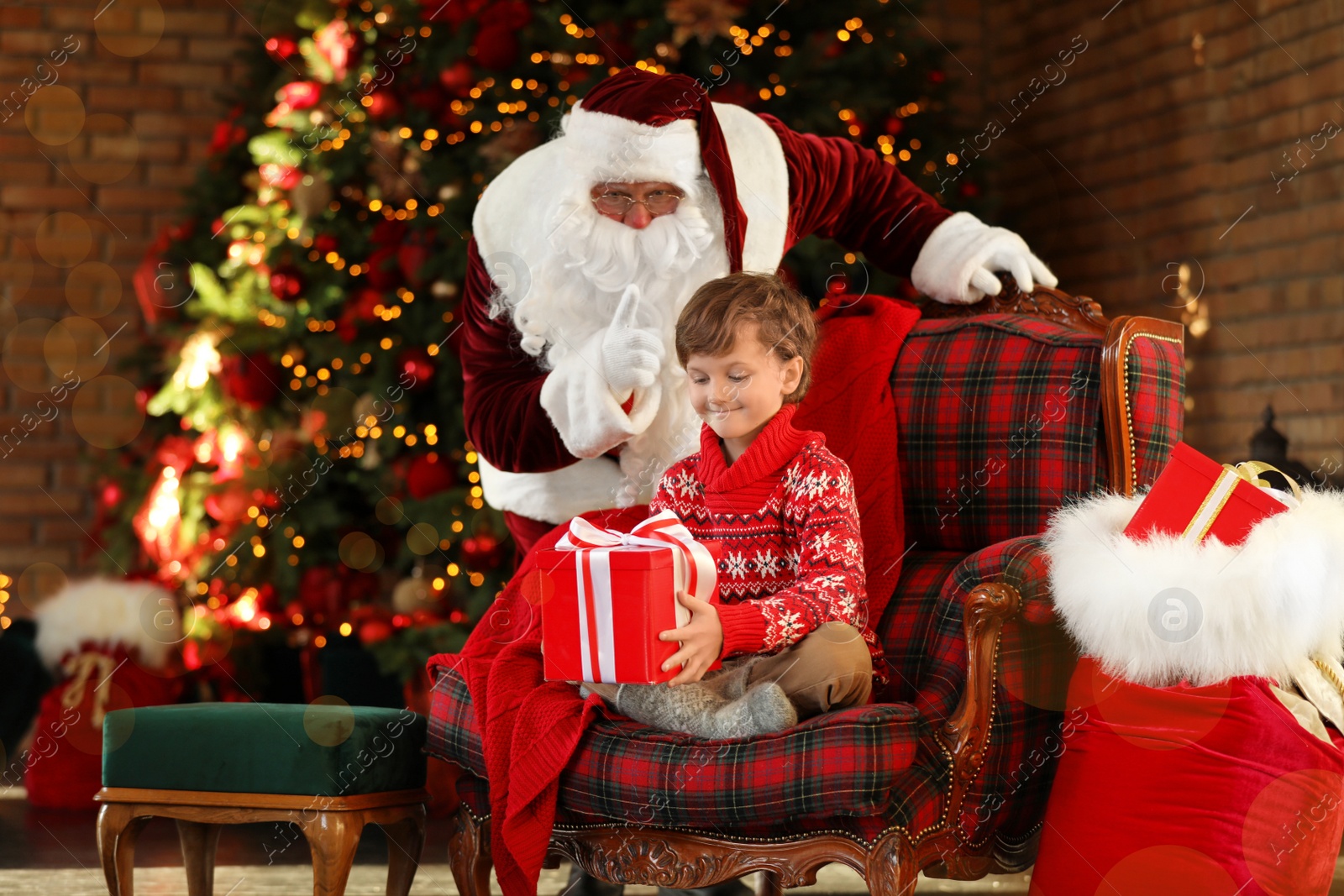 Photo of Santa Claus and little boy with gift near Christmas tree indoors