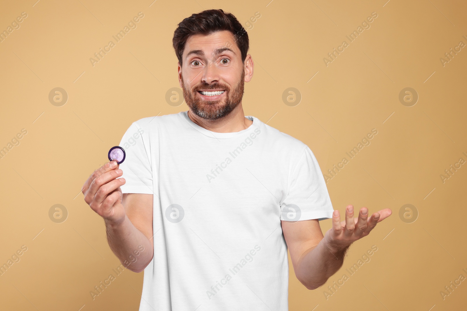 Photo of Confused man holding condom on beige background. Safe sex