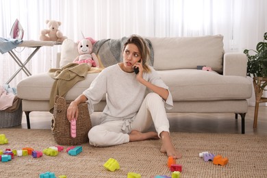 Photo of Tired young mother talking on phone in messy living room