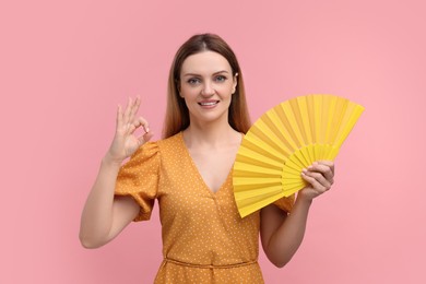 Photo of Happy woman with yellow hand fan showing OK gesture on pink background