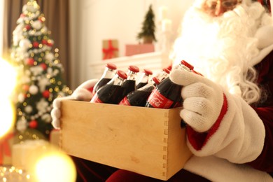 Photo of MYKOLAIV, UKRAINE - JANUARY 18, 2021: Santa Claus holding wooden crate full of Coca-Cola bottles in room with Christmas tree, closeup